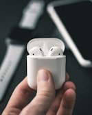 A close-up shot of a hand holding a white wireless earphone case with earphones inside. In the blurred background, a smartwatch and a smartphone are partially visible.