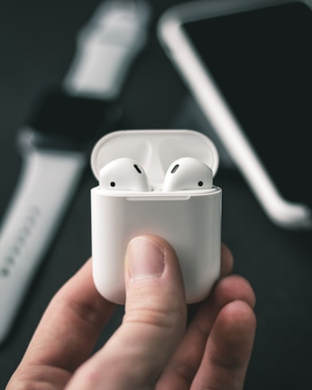 A close-up shot of a hand holding a white wireless earphone case with earphones inside. In the blurred background, a smartwatch and a smartphone are partially visible.