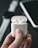 A close-up shot of a hand holding a white wireless earphone case with earphones inside. In the blurred background, a smartwatch and a smartphone are partially visible.
