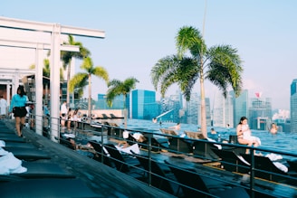 A rooftop infinity pool area overlooking a modern city skyline. There are palm trees, lounge chairs with white towels, and a few people relaxing by the pool. The sky is clear and blue. A person is walking with a tray on the left side of the image.