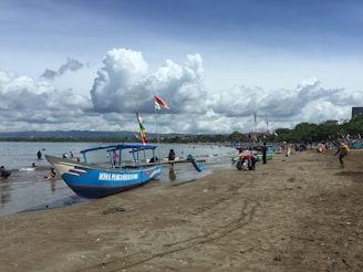 A sandy beach with a large wooden boat painted blue and white, featuring flags and the name 'Mina Panganiban 004'. People are scattered along the shore and in the water, enjoying a sunny day. The background showcases a line of green trees and distant mountains under a sky filled with fluffy clouds.