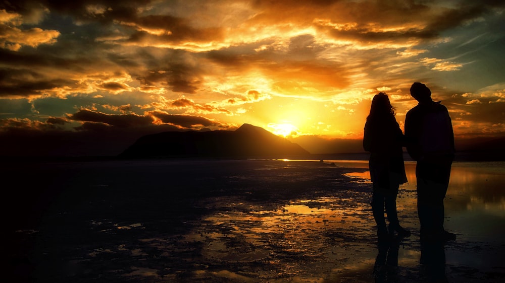 silhouette photography of two person standing on lake during golden hour