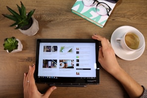 A person is holding a tablet on a wooden table, which displays several articles in a tiled format. To the left, there are two small potted plants. On the upper right, a pair of glasses rests on a stack of books, and to the right is a cup of coffee on a saucer.