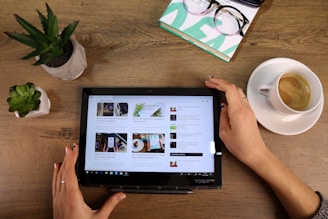 A person is holding a tablet on a wooden table, which displays several articles in a tiled format. To the left, there are two small potted plants. On the upper right, a pair of glasses rests on a stack of books, and to the right is a cup of coffee on a saucer.