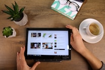 A person is holding a tablet on a wooden table, which displays several articles in a tiled format. To the left, there are two small potted plants. On the upper right, a pair of glasses rests on a stack of books, and to the right is a cup of coffee on a saucer.