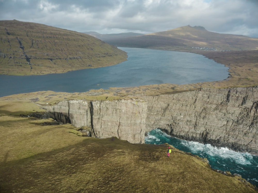 high-angle photography of person standing on brown mountains with body of waters below