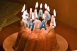 A bundt cake adorned with blue-lettered candles spelling 'Happy Birthday' is set on a white plate. The candles are lit, creating a warm glow against the cake, which is lightly dusted with powdered sugar. Behind the cake is a subtly visible striped gift wrap.