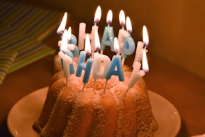 A bundt cake adorned with blue-lettered candles spelling 'Happy Birthday' is set on a white plate. The candles are lit, creating a warm glow against the cake, which is lightly dusted with powdered sugar. Behind the cake is a subtly visible striped gift wrap.