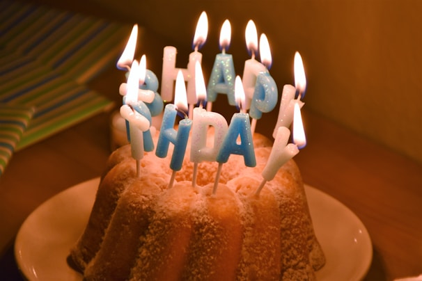 A bundt cake adorned with blue-lettered candles spelling 'Happy Birthday' is set on a white plate. The candles are lit, creating a warm glow against the cake, which is lightly dusted with powdered sugar. Behind the cake is a subtly visible striped gift wrap.
