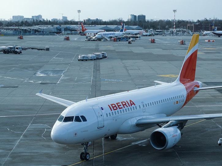 A commercial airliner from the Iberia airline is parked on a tarmac at an airport. Various other planes from different airlines are visible in the background, alongside airport vehicles and scattered cargo. The sky is overcast and the airport buildings are visible in the distance.
