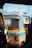 A close-up view of a colorful auto rickshaw featuring a worn and weathered look. Its canopy is a dark shade, covering the back, and the body is yellow with a prominent blue stripe. This image captures an urban scene with visible structure in the background and another part of a vehicle in the foreground.