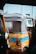 A close-up view of a colorful auto rickshaw featuring a worn and weathered look. Its canopy is a dark shade, covering the back, and the body is yellow with a prominent blue stripe. This image captures an urban scene with visible structure in the background and another part of a vehicle in the foreground.