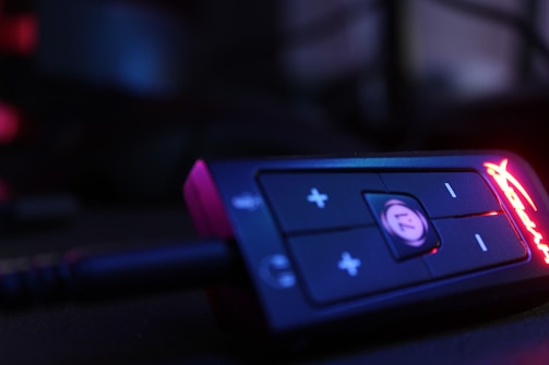 A close-up view of an electronic device showcasing a control panel with buttons and LED lighting. The device is mostly black with prominent plus and minus buttons, and a glowing red brand logo is visible on the right side.