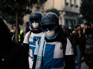 Two individuals wearing helmets, goggles, protective masks, and white shirts with blue crosses are walking in a crowd on a street. Other people in the background appear blurred, contributing to a sense of movement and urgency.