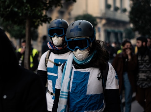 Two individuals wearing helmets, goggles, protective masks, and white shirts with blue crosses are walking in a crowd on a street. Other people in the background appear blurred, contributing to a sense of movement and urgency.