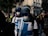 Two individuals wearing helmets, goggles, protective masks, and white shirts with blue crosses are walking in a crowd on a street. Other people in the background appear blurred, contributing to a sense of movement and urgency.