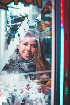 A woman is smiling and looking at a claw machine filled with plush toys. The scene is set indoors with a mix of bright and neon lights reflecting off the glass. She is wearing a scarf and her hair is let down.