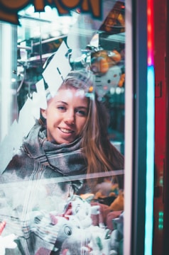 A woman is smiling and looking at a claw machine filled with plush toys. The scene is set indoors with a mix of bright and neon lights reflecting off the glass. She is wearing a scarf and her hair is let down.