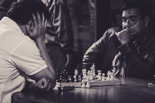 Two individuals are deeply engrossed in a chess game, sitting across from each other at a wooden table. One person appears contemplative, resting his head in one hand, while the other looks focused, with his chin resting on his hand. A smartphone is placed on the table near the chessboard, which is actively in use with several pieces on it.
