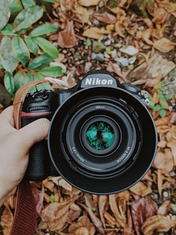 A hand-held Nikon DSLR camera with a lens that reflects green foliage is centered over a background of fallen brown leaves and some green foliage.