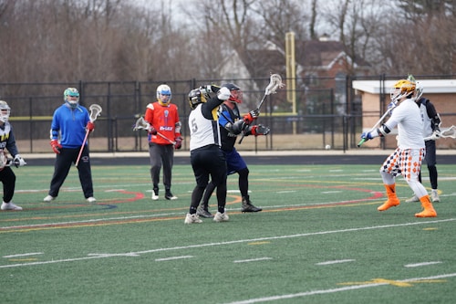 A group of lacrosse players are actively engaged in a practice or game on a sports field. The players are wearing various uniforms and protective gear, including helmets and gloves. The field is marked with multiple colored lines, and there are trees and a building in the background.