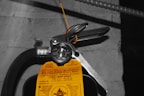 A close-up view of a fire extinguisher with a yellow inspection tag attached. The tag contains details about the service and inspection of the equipment. A pressure gauge is visible, indicating the operational status of the extinguisher. The background is a concrete wall with an electrical conduit running parallel to the extinguisher.