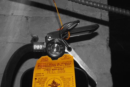 A close-up view of a fire extinguisher with a yellow inspection tag attached. The tag contains details about the service and inspection of the equipment. A pressure gauge is visible, indicating the operational status of the extinguisher. The background is a concrete wall with an electrical conduit running parallel to the extinguisher.