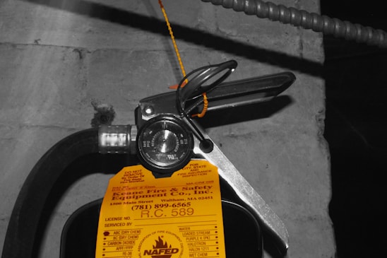 A close-up view of a fire extinguisher with a yellow inspection tag attached. The tag contains details about the service and inspection of the equipment. A pressure gauge is visible, indicating the operational status of the extinguisher. The background is a concrete wall with an electrical conduit running parallel to the extinguisher.