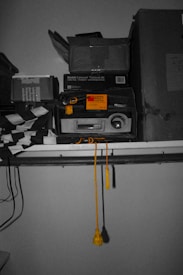 A collection of vintage items stacked on a shelf. There is a Kodak Carousel Transvue 80 slide tray box and a rectangular projector. A bright yellow string with tassels hangs from the shelf, and there are various boxes and packages around.