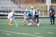 A group of lacrosse players are in action on a sports field. The players are wearing helmets and various athletic gear. One player is attempting to scoop the ball with his stick while others are closely following. The field has markings, and empty bleachers are visible in the background.
