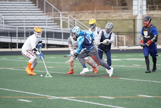A group of lacrosse players are in action on a sports field. The players are wearing helmets and various athletic gear. One player is attempting to scoop the ball with his stick while others are closely following. The field has markings, and empty bleachers are visible in the background.