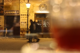 A cozy street scene with warm lighting featuring a vintage-style street lamp illuminating the sidewalk. Two people in winter clothing walk past, while blurred reflections in the foreground create a soft, dreamy effect. Store signs, including 'Laurinská 11' and 'Roxy Music Shop,' are visible, and snow covers the ground.