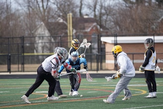 A group of lacrosse players are actively engaged in a game on a field with lines and markings in yellow, red, and white. The players are wearing protective gear, including helmets and gloves, with some in jerseys labeled with numbers and team names. Bare trees and a building can be seen in the background beyond a chain-link fence.