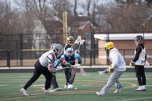 A group of lacrosse players are actively engaged in a game on a field with lines and markings in yellow, red, and white. The players are wearing protective gear, including helmets and gloves, with some in jerseys labeled with numbers and team names. Bare trees and a building can be seen in the background beyond a chain-link fence.