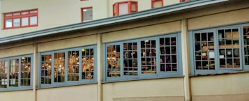 A building facade featuring large, multi-pane windows with visible interior lights. The setting appears to be a street or alley with part of the building shown at an angle, suggesting an urban environment. Inside the windows, various items and furniture are visible, indicating a possible shop or showroom.