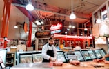 A butcher stands behind a wooden counter in a bustling market environment. The interior is warmly lit with exposed piping and a neon sign overhead advertising Market Spice. Various cuts of meat are displayed in refrigerated cases, and the space is vibrant with activity.
