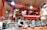 A butcher stands behind a wooden counter in a bustling market environment. The interior is warmly lit with exposed piping and a neon sign overhead advertising Market Spice. Various cuts of meat are displayed in refrigerated cases, and the space is vibrant with activity.