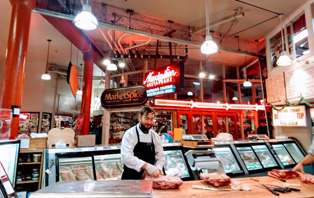 A butcher stands behind a wooden counter in a bustling market environment. The interior is warmly lit with exposed piping and a neon sign overhead advertising Market Spice. Various cuts of meat are displayed in refrigerated cases, and the space is vibrant with activity.