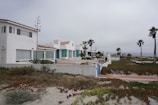 A row of white, modern beach houses with tile roofs situated along a sandy coast. The weather is overcast with a grey sky. In the foreground, there is a mix of coastal plants and sand. Palm trees are scattered along the landscape, and a walkway runs parallel to the houses.