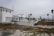 A row of white, modern beach houses with tile roofs situated along a sandy coast. The weather is overcast with a grey sky. In the foreground, there is a mix of coastal plants and sand. Palm trees are scattered along the landscape, and a walkway runs parallel to the houses.