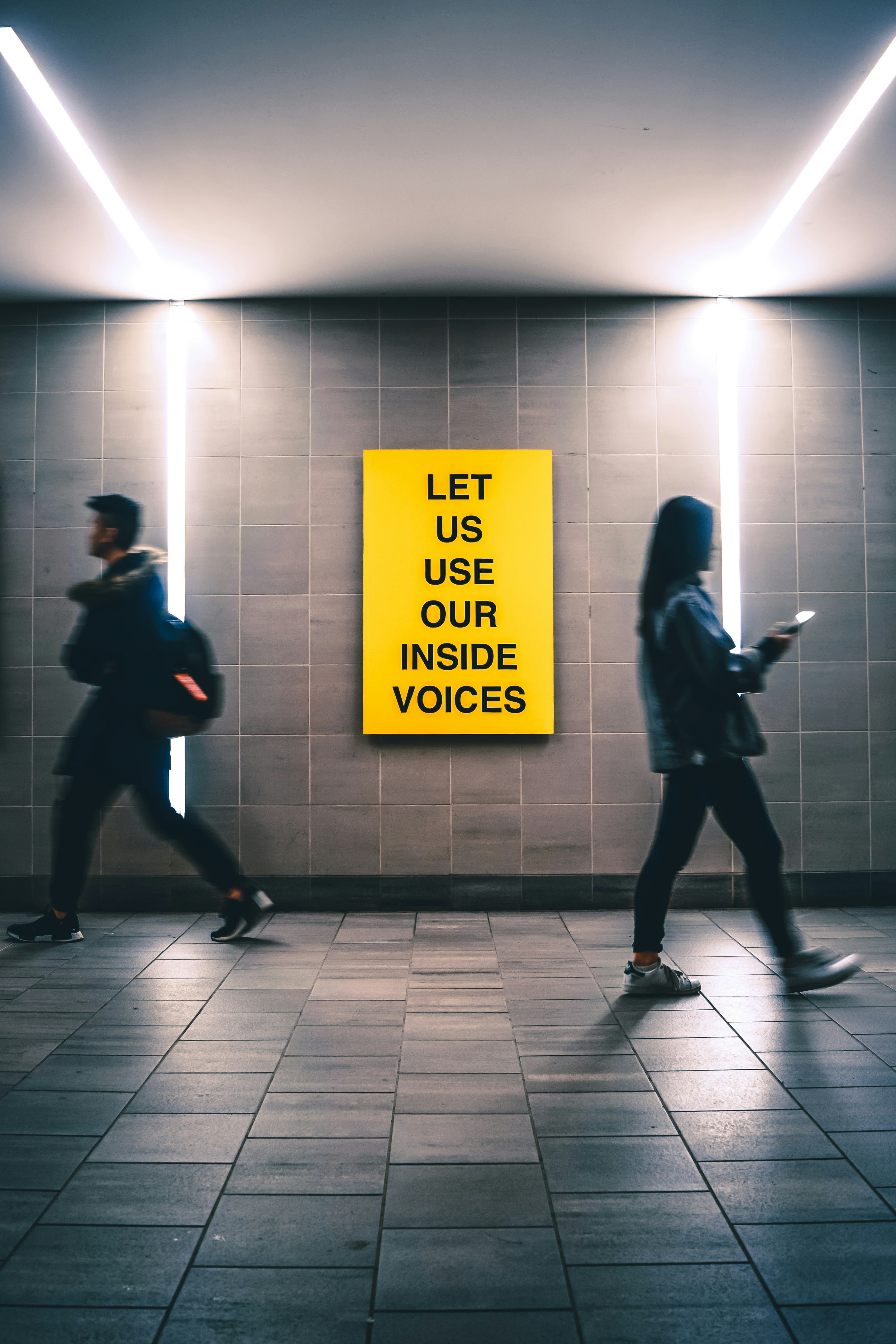 man and woman walking inside building near Let us use our inside voices sign