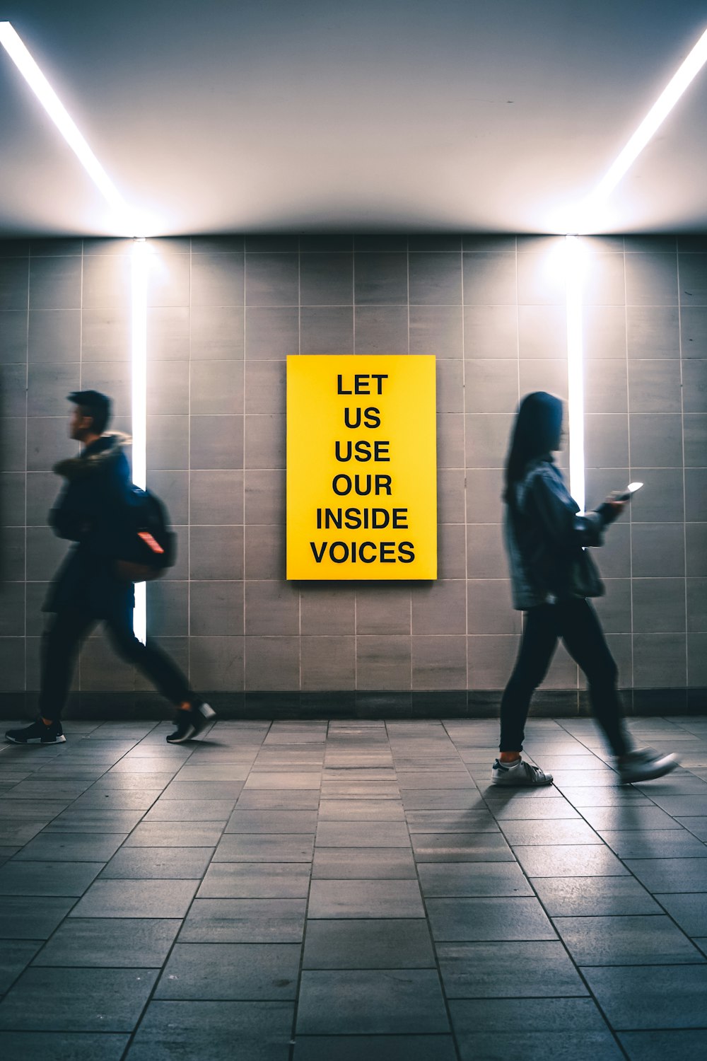 man and woman walking inside building near Let us use our inside voices sign