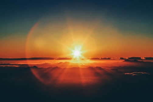 A vibrant sunrise with the sun partially visible over a range of silhouetted mountains. The sky is filled with warm hues of orange and red, and gentle clouds float above the mountain peaks, reflecting the morning light.