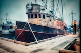 A large fishing boat is docked at a snowy pier. The boat appears weathered, with metal structures and ropes prominent. Snow is gently falling, creating a serene winter scene. The pier is covered in a light dusting of snow, and a small lamp illuminates the area.