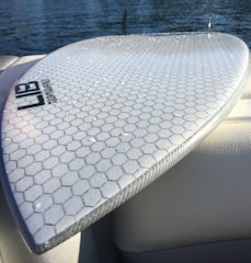 A surfboard with a hexagonal pattern on its surface is resting on a boat seat near a body of water. Water droplets are visible on the board, and part of the lake or sea can be seen in the background.