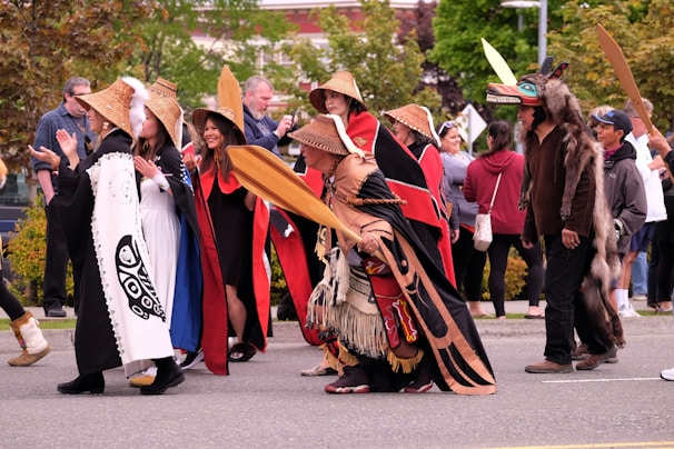 A group of people participates in a cultural parade or gathering, wearing traditional clothing and carrying wooden paddles. The attire includes woven hats and garments with intricate designs. There are spectators in the background observing the event, with some people capturing the moment on their phones.
