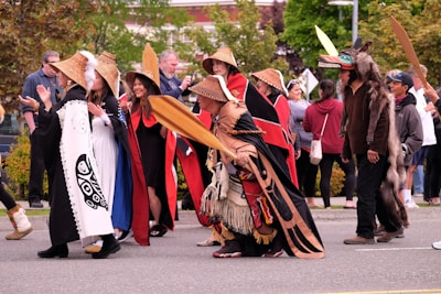 A group of people participates in a cultural parade or gathering, wearing traditional clothing and carrying wooden paddles. The attire includes woven hats and garments with intricate designs. There are spectators in the background observing the event, with some people capturing the moment on their phones.
