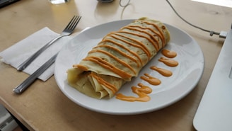 A rolled crepe topped with a zigzag pattern of orange sauce is placed on a white plate. The plate is on a wooden surface with a napkin, fork, and knife beside it. Part of a laptop and a glass of water are visible in the background.