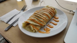 A rolled crepe topped with a zigzag pattern of orange sauce is placed on a white plate. The plate is on a wooden surface with a napkin, fork, and knife beside it. Part of a laptop and a glass of water are visible in the background.