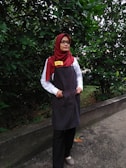A person is standing outdoors in front of dense green foliage. They are wearing a maroon headscarf, glasses, a white shirt, a dark apron, and black pants. The person has a name tag that reads 'DINIE'. The background consists of a lush green hedge and a concrete path.